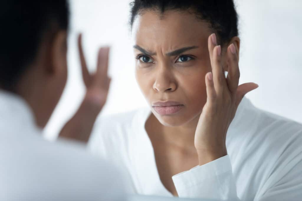 Woman looking in the mirror ACat facial wrinkles and considering BOTOX vs. dermal fillers